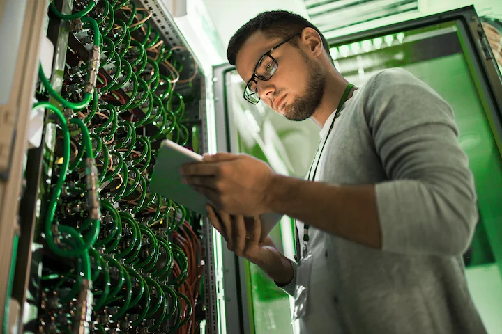 Person looking at a laptop standing at a network rack